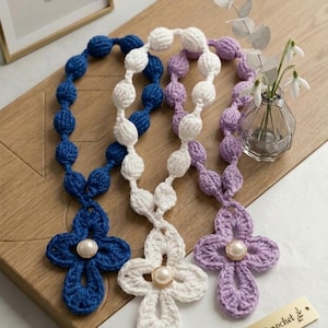 May include: Three handmade crochet rosaries in blue, white, and lavender. Each rosary features a cross pendant with a pearl-like bead at the center. The rosaries are displayed on a wooden surface, with a small vase of flowers in the background.