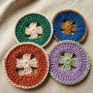 K&ouml;nnte beinhalten: Vier handgefertigte H&auml;keluntersetzer in Gr&uuml;n, Blau, Orange und Lila. Jeder Untersetzer hat ein zentrales Kreuzdesign und einen kontrastierenden Rand. Die runden Untersetzer liegen auf einer neutralen Stoffoberfl&auml;che.