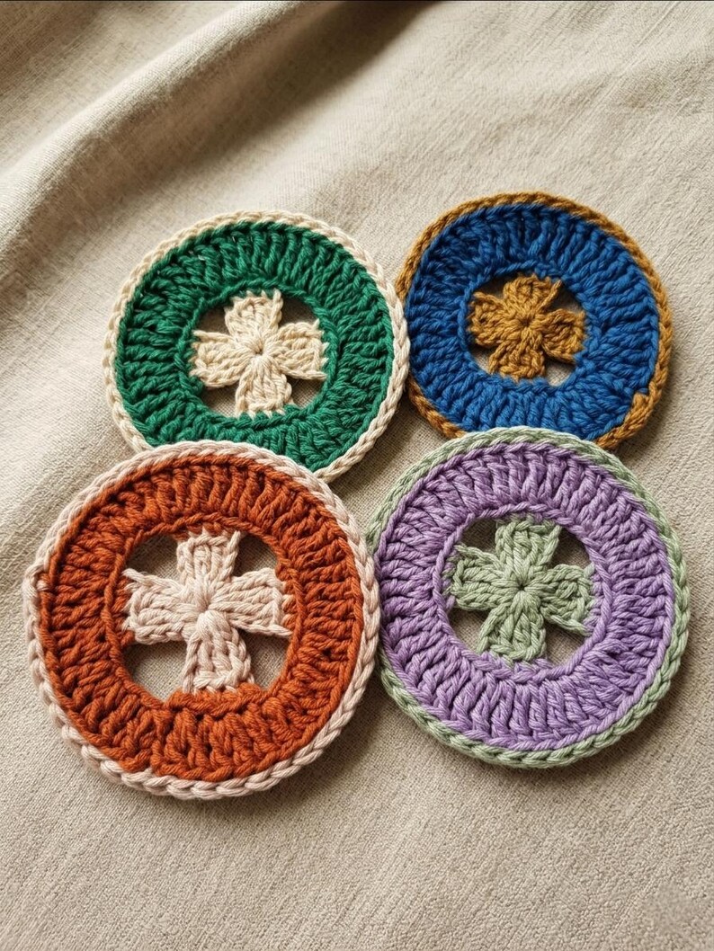 K&ouml;nnte beinhalten: Vier handgefertigte H&auml;keluntersetzer in Gr&uuml;n, Blau, Orange und Lila. Jeder Untersetzer hat ein zentrales Kreuzdesign und einen kontrastierenden Rand. Die runden Untersetzer liegen auf einer neutralen Stoffoberfl&auml;che.
