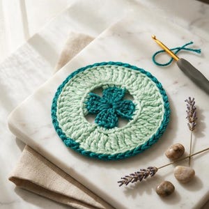 K&ouml;nnte beinhalten: Handgefertigter H&auml;keluntersetzer in T&uuml;rkis und Mintgr&uuml;n mit einem vierbl&auml;ttrigen Kleeblatt-Design in der Mitte. Der Untersetzer liegt auf einer Marmoroberfl&auml;che, daneben eine H&auml;kelnadel und Lavendelzweige. Ein Bastelprojekt.