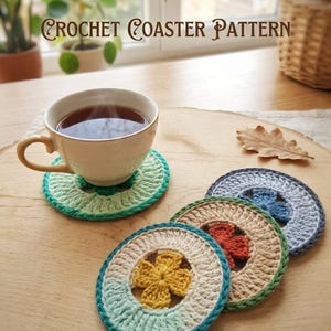 K&ouml;nnte beinhalten: Eine Tasse Tee steht auf einem gr&uuml;nen geh&auml;kelten Untersetzer, mit mehreren anderen bunten Untersetzern in der N&auml;he. Die Untersetzer sind kreisf&ouml;rmig mit einem Blumenmuster in der Mitte. Der Text "GOKKUSAGICROCHET CROCHET COASTER PATTERN" ist sichtbar.