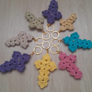 May include: A collection of handmade crochet cross keychains in various colors, including purple, yellow, pink, and teal. Each cross features a gold-colored metal keyring and a small gold bead in the center. The keychains are arranged on a light wooden surface.