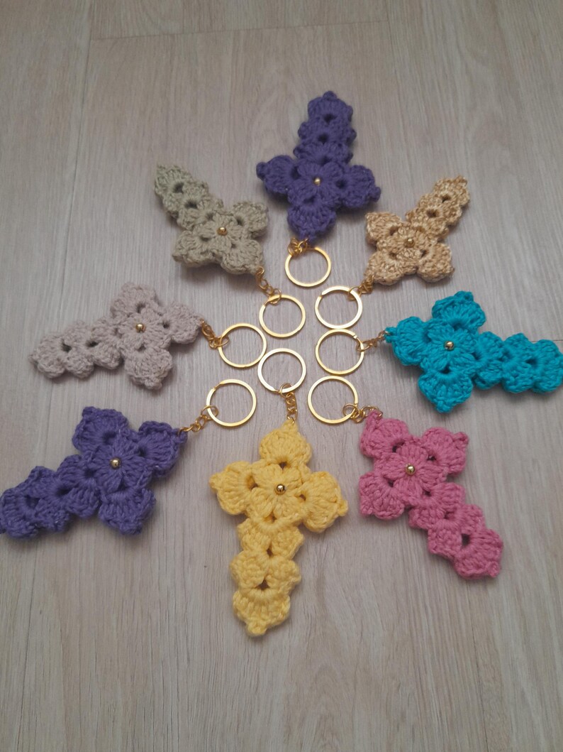 May include: A collection of handmade crochet cross keychains in various colors, including purple, yellow, pink, and teal. Each cross features a gold-colored metal keyring and a small gold bead in the center. The keychains are arranged on a light wooden surface.