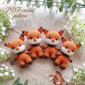 May include: Four crocheted fox toys in orange, white, and brown, arranged on a wooden surface with greenery. The foxes have detailed features. The image includes the text "PDF crochet pattern".
