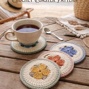 K&ouml;nnte beinhalten: Handgefertigte H&auml;keluntersetzer in Creme, Blau, Orange und Gelb mit floralen Designs, auf einem Holztisch angeordnet. Eine Tasse Tee steht auf einem Untersetzer. Der Text "CROCHET COASTER PATTERN" ist im Bild zu sehen.