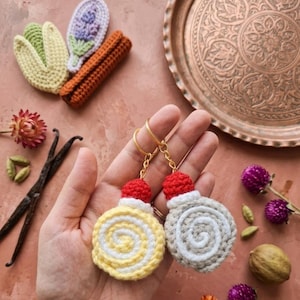 May include: Two round, crocheted keychains featuring a spiral pattern in yellow and white or beige and white, each topped with a small red crocheted sphere and a gold-toned metal keyring, showcasing intricate yarn textures and craftsmanship.