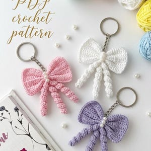 May include: Handmade crochet bow keyrings in pink, white, and purple. Each bow has a pearl accent and twisted cord details. The image includes the text "PDF crochet pattern" and several balls of yarn.
