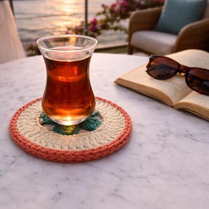 K&ouml;nnte beinhalten: Ein Glas Tee steht auf einem handgefertigten H&auml;keluntersetzer in Pfirsich-, Creme- und T&uuml;rkist&ouml;nen. Sonnenbrille und ein offenes Buch liegen in der N&auml;he auf einem Marmortisch. Der Hintergrund zeigt einen Sonnenuntergang &uuml;ber dem Wasser.