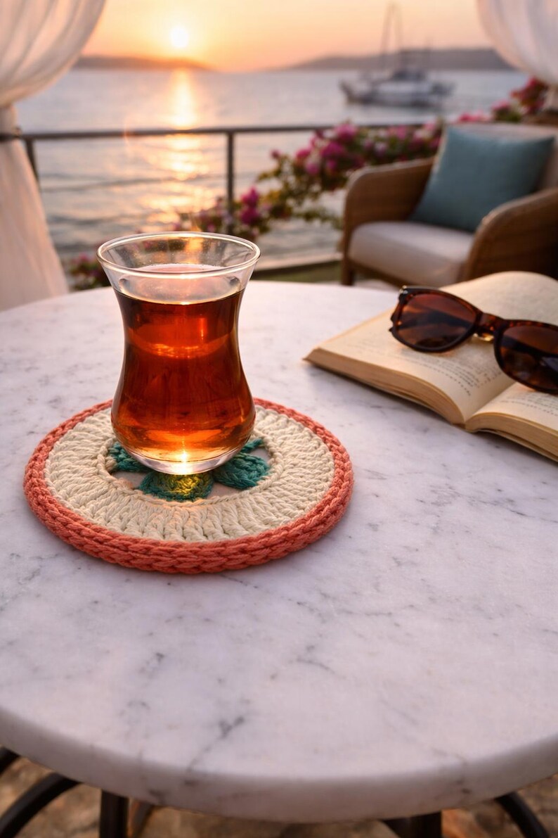 K&ouml;nnte beinhalten: Ein Glas Tee steht auf einem handgefertigten H&auml;keluntersetzer in Pfirsich-, Creme- und T&uuml;rkist&ouml;nen. Sonnenbrille und ein offenes Buch liegen in der N&auml;he auf einem Marmortisch. Der Hintergrund zeigt einen Sonnenuntergang &uuml;ber dem Wasser.