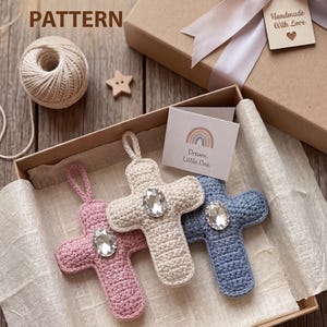 Puede incluir: Tres cruces de ganchillo en rosa, crema y azul, cada una con una gran gema brillante en el centro, se exhiben en una caja de regalo. La caja está atada con una cinta y una etiqueta que dice "Handmade With Love". El texto "PDF CROCHET PATTERN" está en la parte superior.