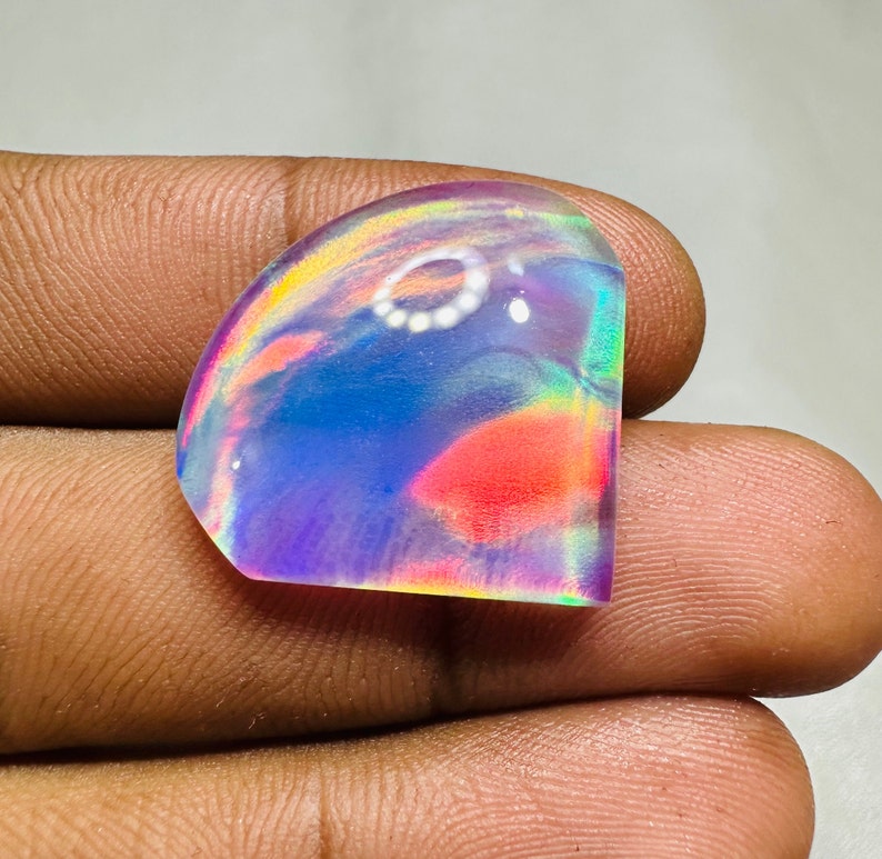 May include: A cabochon cut iridescent opal gemstone with a rainbow of colors, including pink, blue, green, and purple. The opal is set in a clear, faceted setting.