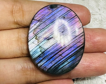 May include: Oval-shaped labradorite gemstone with a polished surface. The stone displays a vibrant play of color, with hues of purple, blue, and pink. Diagonal lines create a striped pattern across the surface, enhancing the stone's visual appeal. The gemstone is held in a hand.