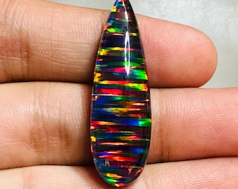 May include: A polished, teardrop-shaped black opal gemstone. The opal displays vibrant bands of color, including red, green, and yellow, creating a striking play of light. The gemstone is held in a hand.