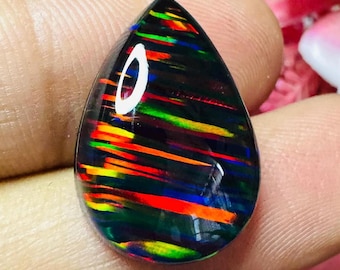 May include: A teardrop-shaped black opal gemstone with vibrant streaks of red, orange, yellow, and green. The polished surface reflects light, showcasing the play of color within the stone. The opal is held in a hand.