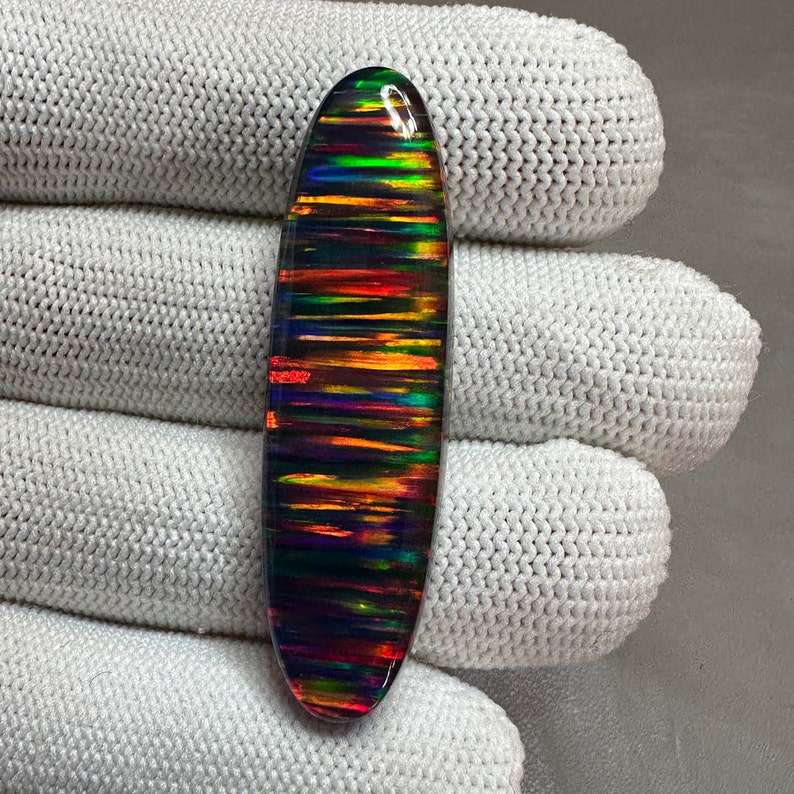 May include: A colorful, oval-shaped opal gemstone with a black background and iridescent stripes of red, orange, yellow, green, blue, and purple.