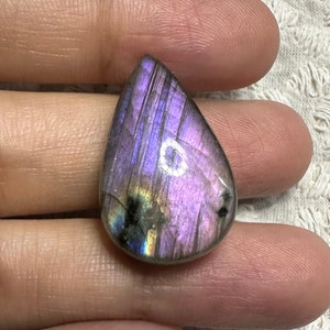 May include: A teardrop-shaped, iridescent labradorite gemstone with a purple, blue, and green sheen. The stone is polished and has a smooth surface.