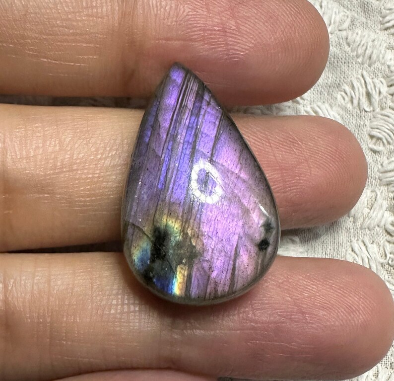 May include: A teardrop-shaped, iridescent labradorite gemstone with a purple, blue, and green sheen. The stone is polished and has a smooth surface.