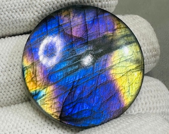 May include: A round, iridescent labradorite gemstone cabochon with a blue, purple, and yellow color pattern. The stone is polished and has a smooth, shiny surface.