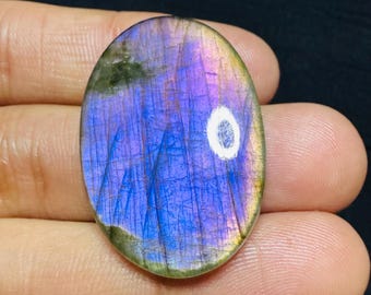 Op de afbeelding: Ovale labradoriet edelsteen met een levendige weergave van blauw, paars en groen iriserend. De gepolijste steen heeft een glad oppervlak en een witte insluitsel in het midden. De edelsteen wordt in een hand gehouden.