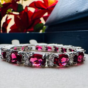 May include: A silver-toned link bracelet featuring alternating oval-cut vibrant red gemstones and clusters of small, clear, round-cut white stones. The bracelet is arranged in a repeating pattern of red stones separated by pairs of white stones.