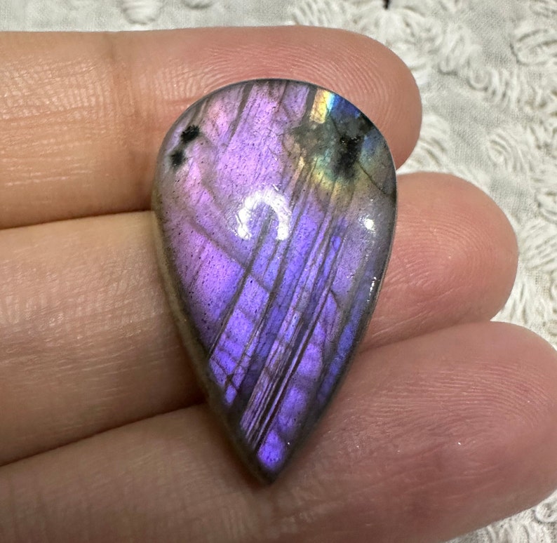 May include: A teardrop-shaped cabochon of labradorite gemstone with iridescent purple, blue, and green hues.