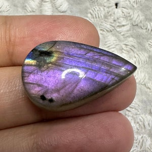 May include: A teardrop-shaped, iridescent labradorite gemstone with purple, blue, and green hues. The stone has a smooth, polished surface and is set in a cabochon cut.