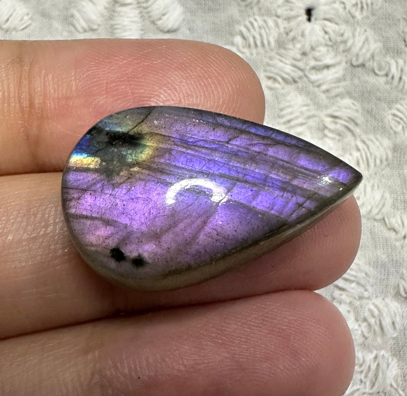 May include: A teardrop-shaped, iridescent labradorite gemstone with purple, blue, and green hues. The stone has a smooth, polished surface and is set in a cabochon cut.