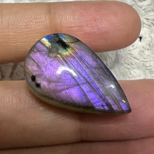 May include: A teardrop-shaped, iridescent labradorite gemstone with a purple and gray color scheme. The stone has a smooth, polished surface and displays a rainbow of colors when light shines through it.