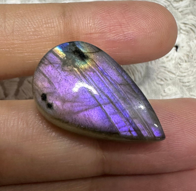 May include: A teardrop-shaped, iridescent labradorite gemstone with a purple and gray color scheme. The stone has a smooth, polished surface and displays a rainbow of colors when light shines through it.