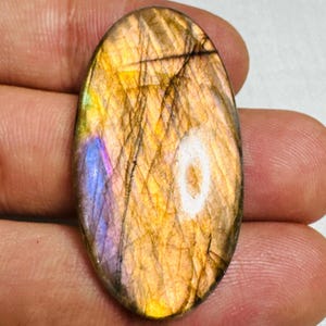 May include: Oval-shaped labradorite gemstone with a polished surface. The stone displays a mix of colors, including gold, brown, purple, and blue, with a shimmering effect. The gemstone has a network of fine lines across its surface.