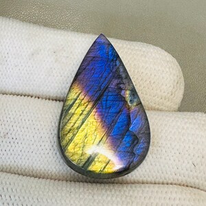 K&ouml;nnte beinhalten: Ein tropfenf&ouml;rmiger Labradorit-Cabochon mit schillernden blauen, violetten und gelben Farben.