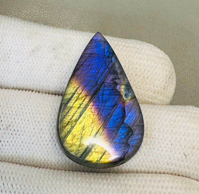 K&ouml;nnte beinhalten: Ein tropfenf&ouml;rmiger Labradorit-Cabochon mit schillernden blauen, violetten und gelben Farben.