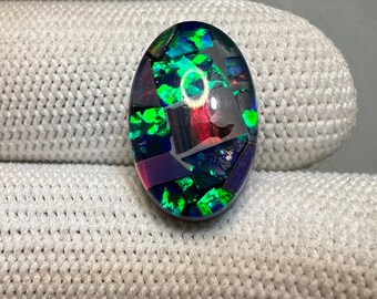 May include: Oval-shaped, iridescent opal cabochon with a mosaic pattern of green, blue, purple, and red. The opal has a high shine and is set in a clear resin.