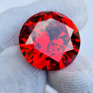 Puede incluir: Una gema roja grande, facetada y de corte redondo. La gema tiene muchas superficies reflectantes, creando una brillante exhibición de luz y color. La gema es sostenida por un guante blanco.