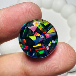May include: A circular cabochon featuring a mosaic of iridescent opal fragments set in a dark, black base. The surface displays a vibrant array of multicolored flashes including red, green, yellow, and blue, arranged in geometric triangular shapes.