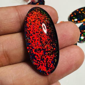 May include: An oval-shaped gemstone with a black base and a vibrant display of red, orange, and blue internal fire. The gemstone is held in a hand, with other similar stones visible in the background. The stone's surface reflects light.