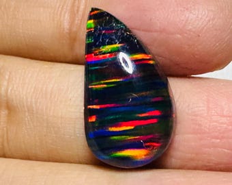 May include: A teardrop-shaped black opal gemstone with vibrant horizontal bands of red, orange, yellow, and green. The polished surface reflects light, showcasing the stone's internal play of color. The opal is held between fingers.