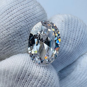 May include: A large, oval-cut diamond with numerous facets, reflecting light in a spectrum of colors. The gemstone is held against a white fabric background, highlighting its brilliance and clarity. The diamond's shape and cut are designed to maximize sparkle.