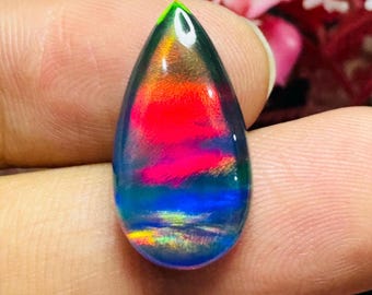 May include: A teardrop-shaped gemstone with a vibrant display of colors. The gem features a gradient of hues, including green, red, blue, and yellow. The stone is held between fingers, showcasing its polished surface and internal color play. This gemstone is suitable for jewelry.