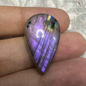 May include: A teardrop-shaped, iridescent labradorite gemstone with purple, blue, and green hues. The stone is polished and has a smooth, glossy finish.