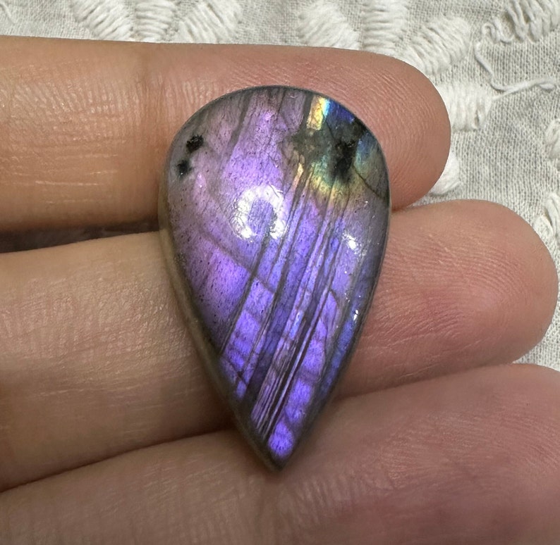 May include: A teardrop-shaped, iridescent labradorite gemstone with purple, blue, and green hues. The stone is polished and has a smooth, glossy finish.