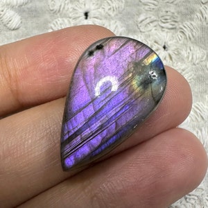 May include: A teardrop-shaped, iridescent labradorite gemstone with purple, gray, and gold hues. The stone is smooth and polished, with visible striations.