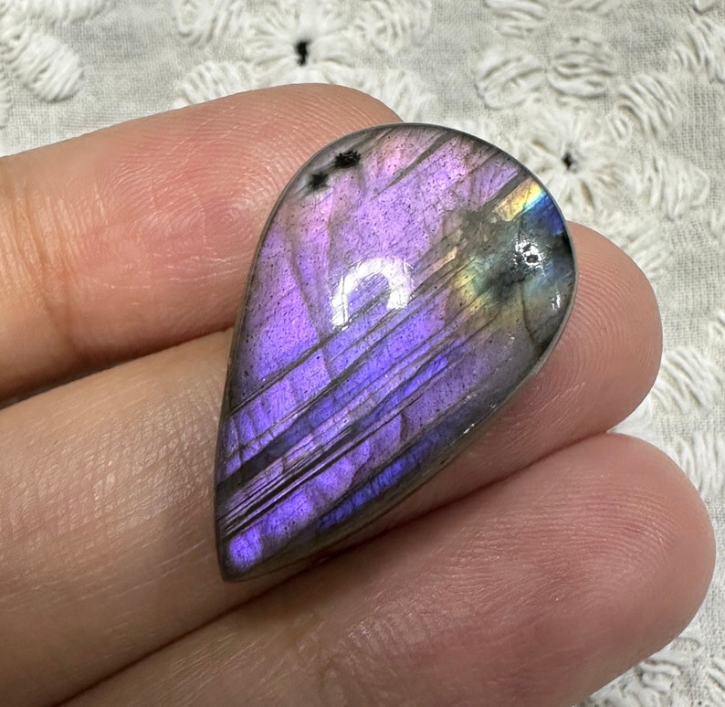May include: A teardrop-shaped, iridescent labradorite gemstone with purple, gray, and gold hues. The stone is smooth and polished, with visible striations.