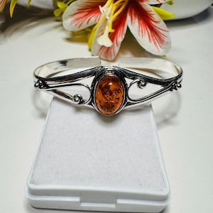 May include: A silver-toned bracelet featuring an oval amber stone at its center. The bracelet has an open design with decorative swirls and curves. The amber stone is a warm, translucent orange color. The bracelet is displayed on a white surface.