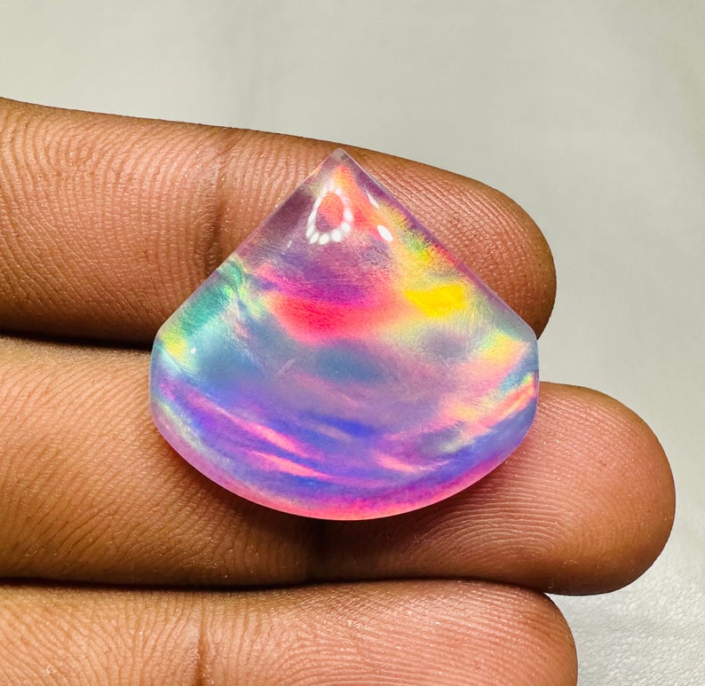 May include: A teardrop-shaped iridescent opal cabochon with a rainbow of colors, including pink, blue, green, and yellow.