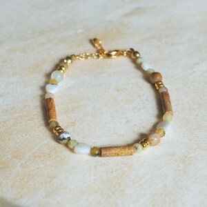 May include: A bracelet with a mix of natural stone beads in brown, white, and yellow. It features cylindrical and round beads, with gold-coloured metal accents and a delicate chain clasp. The bracelet is approximately 18 cm in length.
