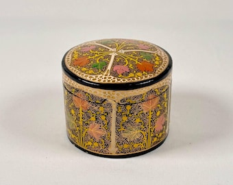 Pode incluir: Uma pequena caixa decorativa redonda, preta e dourada com intrincados desenhos florais em rosa, laranja e verde. A caixa é feita de papel maché e tem uma tampa com dobradiças.