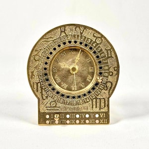 May include: A gold-toned metal clock with a circular face and Roman numeral hour markers. The clock face also features a calendar with numbers 1 through 31. The clock is on a stand with Roman numeral hour markers.