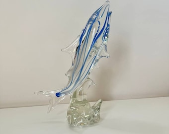 Könnte beinhalten: Eine transparente Glasskulptur eines Fisches mit blauen Streifen, der aus dem Wasser springt. Der Fisch steht auf einem transparenten Glassockel.