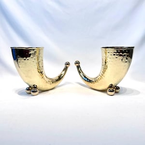 Puede incluir: Dos jarrones decorativos dorados en forma de cuerno. Cada jarrón tiene una textura martillada y descansa sobre tres pequeños pies de bola. Los cuernos se curvan hacia arriba, con una pequeña bola en la punta. Los jarrones se muestran sobre un fondo blanco.
